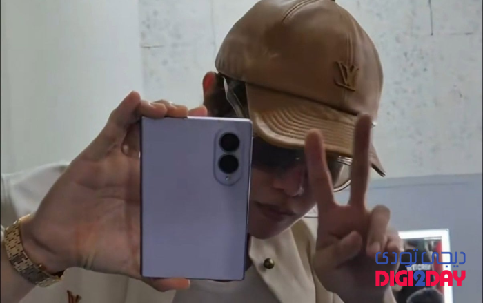 Person takes a mirror selfie with a blue smartphone, wearing a tan cap and glasses, flashing a peace sign with two fingers.