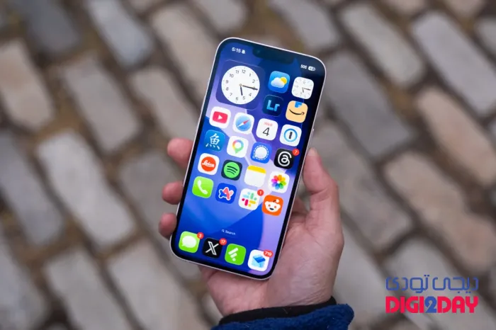 Hand holding a smartphone showing a colorful app grid on the home screen over a cobblestone background.